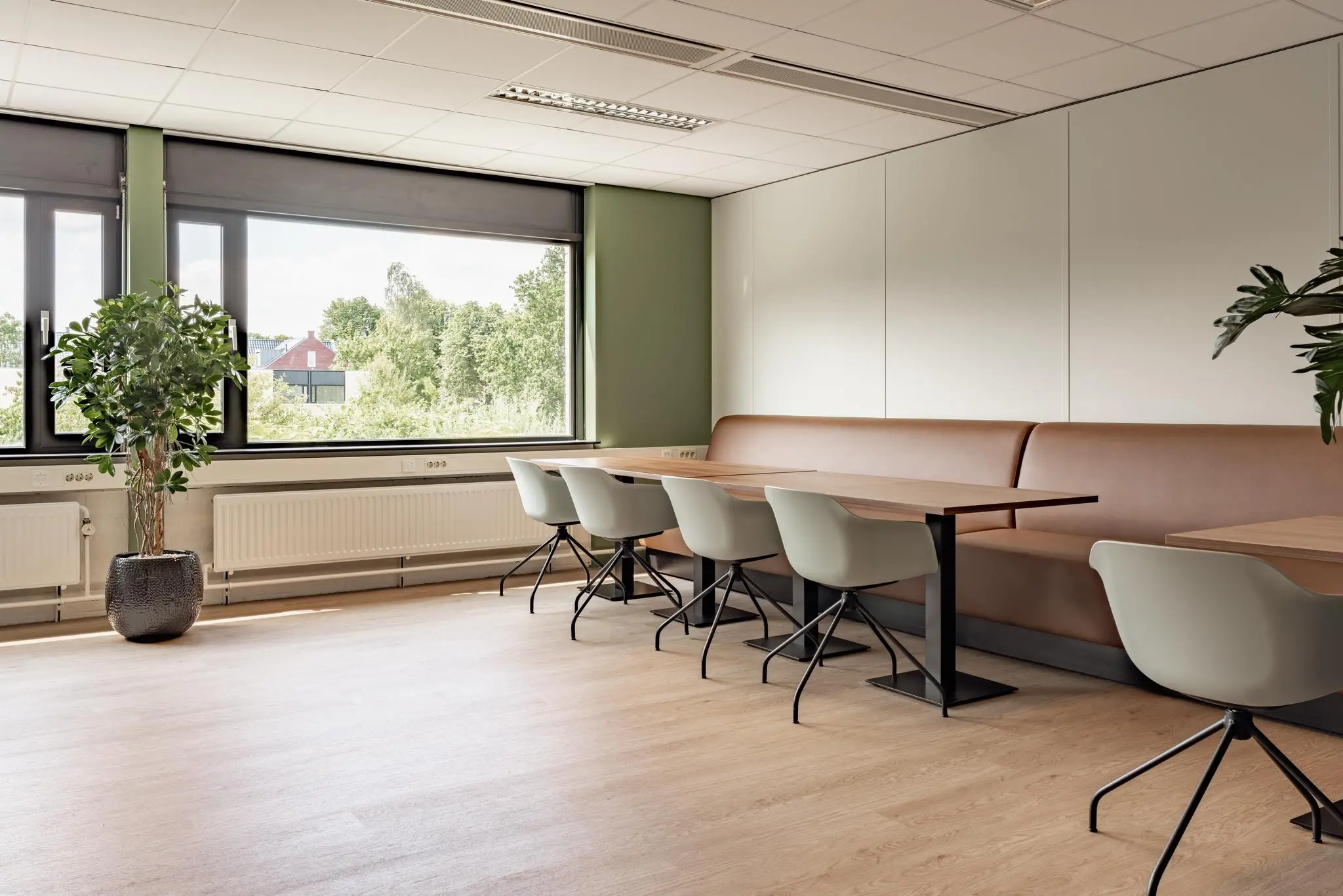 Moderne, lichte zithoek met houten tafels, grijze stoelen en een lange bruine bank in een ruimte aan de Burgemeester Verderlaan.